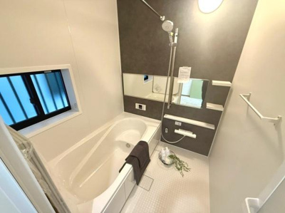 窓があり換気や採光に配慮されたバスルームです。ゆったりとした浴槽で快適に使用できます。落ち着いた色合いの壁面パネルが空間に調和を与えています。収納棚や大きめのミラーも備わっています。