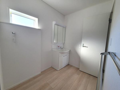 シンプルで明るい洗面台が設置されています。ナチュラルな木目調の床が空間に調和しています。窓から自然光が差し込み清潔感があります。収納スペースも確保されています。壁面もすっきりとした印象です。