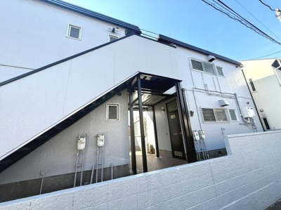 外観は白を基調としたシンプルなデザインです。階段下にはしっかりとした屋根が設けられています。玄関アプローチは明るく共用部分も清潔感があります。電気メーターや設備関係も整理されています。