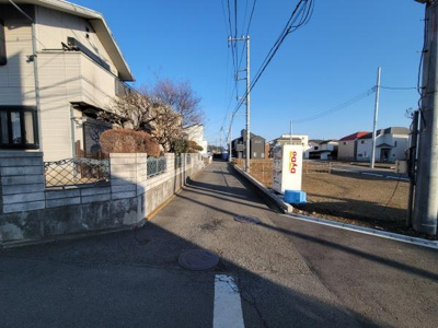 前面道路は広々としており車の出入りがしやすい環境です。周囲には住宅が立ち並び落ち着いた印象を受けます。電柱や歩道も整備されており生活の動線がスムーズです。日差しが十分に差し込む明るい現地写真です。