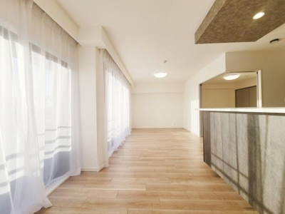 大きな窓から光が差し込む明るく広々としたリビングです。床材の色合いが空間にやわらかな印象を与えます。天井部分にはアクセントとなるデザインが施されています。キッチンカウンターもリビングに調和しています。