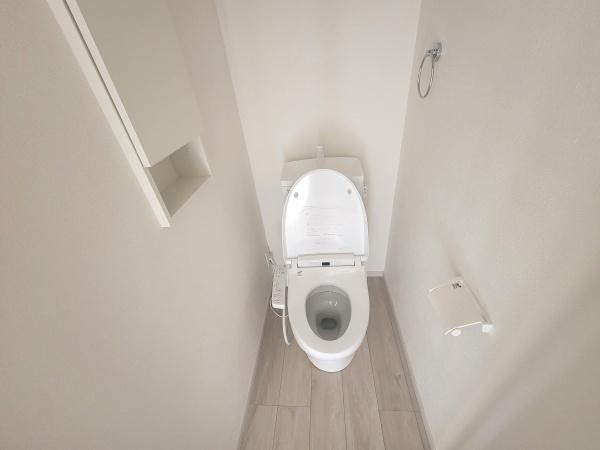 白を基調としたトイレは清潔感があります。壁面の収納スペースも便利です。床材の色味が空間に明るさをプラスしています。ペーパーホルダーやタオルリングも設置されています。