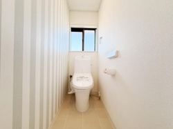 シンプルなデザインのトイレ空間です。窓から自然光が入るため明るい印象です。ストライプ柄の壁がアクセントになっています。タオル掛けやペーパーホルダーも設置されています。清潔感のある仕上がりです。