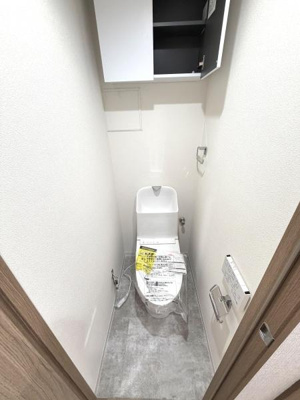 トイレには収納棚が設置されており整理整頓がしやすい空間です。床材が落ち着いた色合いで清潔感があります。ドアや壁面も明るいカラーでシンプルなデザインが特徴です。