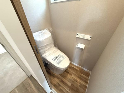 ナチュラルな木目調の床が印象的なトイレです。壁付けのリモコンやペーパーホルダーが設置されています。シンプルな設計で清潔感が感じられます。窓があり換気もしやすい空間です。