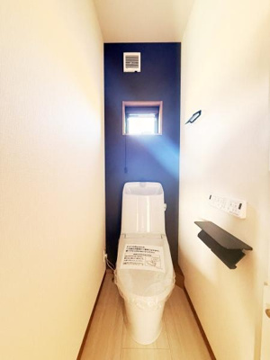 シンプルなデザインのトイレ空間です。明るい内装が清潔感を与えています。小窓から自然光が差し込みます。壁際には収納棚も設置されています。温水洗浄便座が備わっています。