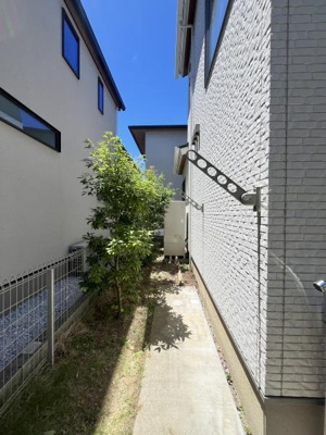 直線的な小道が家の側面を通りシンプルで手入れしやすい外構です。隣家との間にも程よいスペースがあります。外壁の明るい色合いが清潔感を感じさせます。緑の植栽がアクセントとなっています。