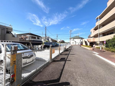 整備された舗装道路と広々とした歩道が印象的な一枚です。周辺は落ち着いた雰囲気が漂い明るい空が開放感を感じさせます。きれいなフェンスと駐車スペースも確認できます。日当たりの良い環境が魅力的に映ります。