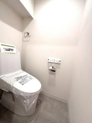 シンプルで清潔感のあるトイレ空間です。壁リモコン設置で操作がしやすい設計となっています。床と壁の色合いが落ち着いた雰囲気を演出します。空間が広めで余裕を感じられます。収納スペースも確保されています。